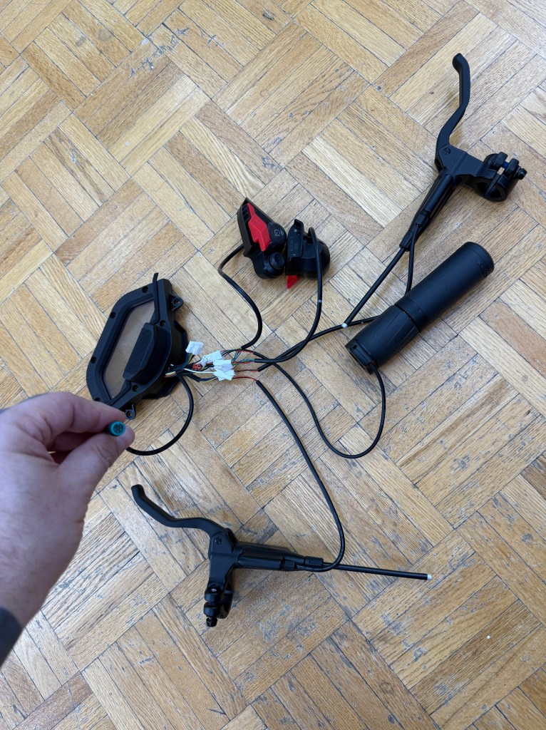 Disassembled bicycle handlebar components including grips, brake levers, and wiring on a wooden floor.