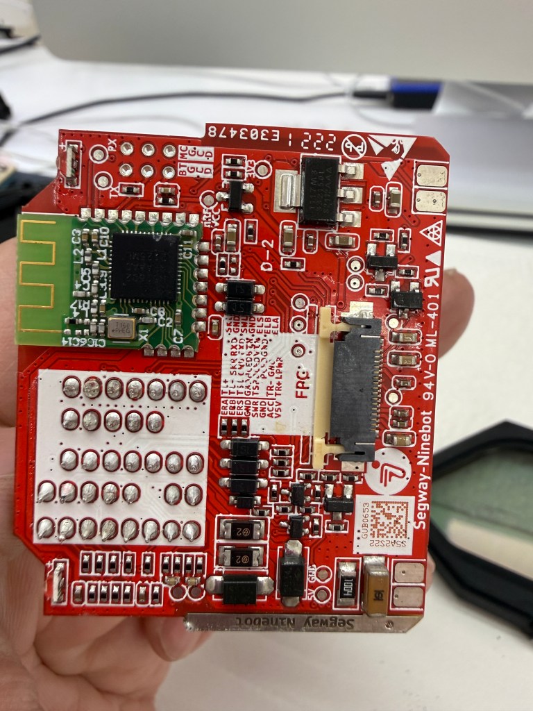 Close-up of a red circuit board with various electronic components, connectors, and printed labels, held in a person's hand.