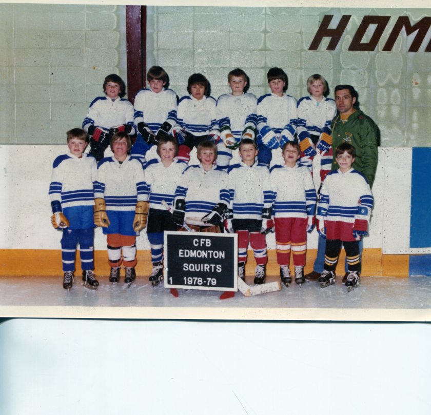 A youth hockey team, the CFB Edmonton Squirts, posing on the ice rink in 1978-79. The team is dressed in white jerseys with blue stripes and red pants, holding hockey sticks, with a coach in a green jacket.